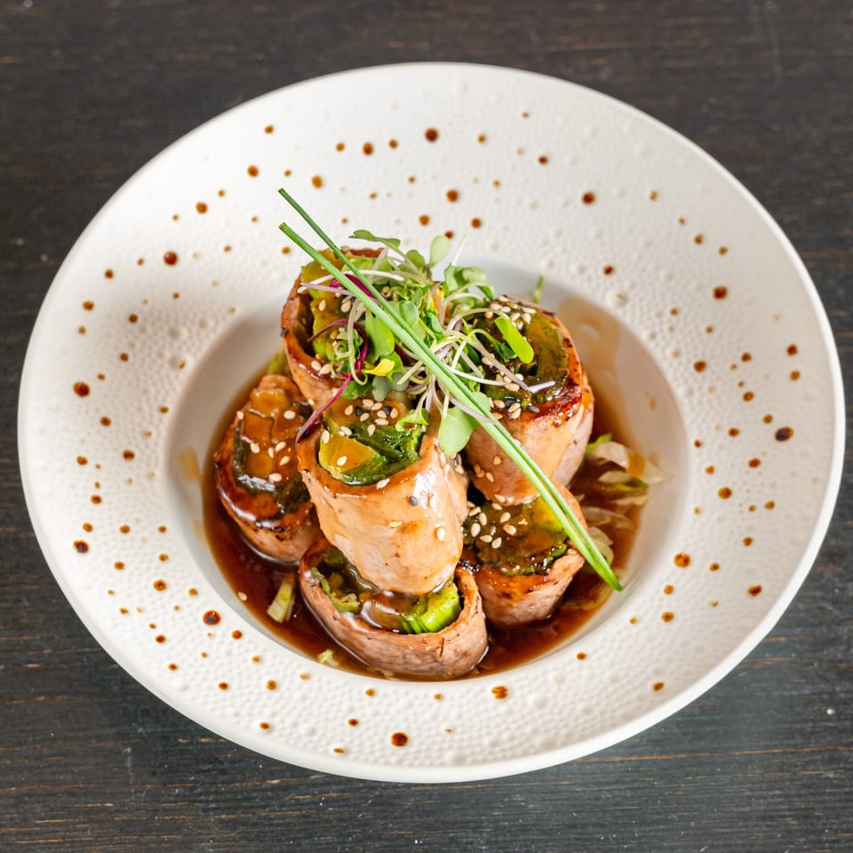 Seared vegetable-filled meat roulades topped with microgreens and sesame seeds in a rich sauce on a white plate.