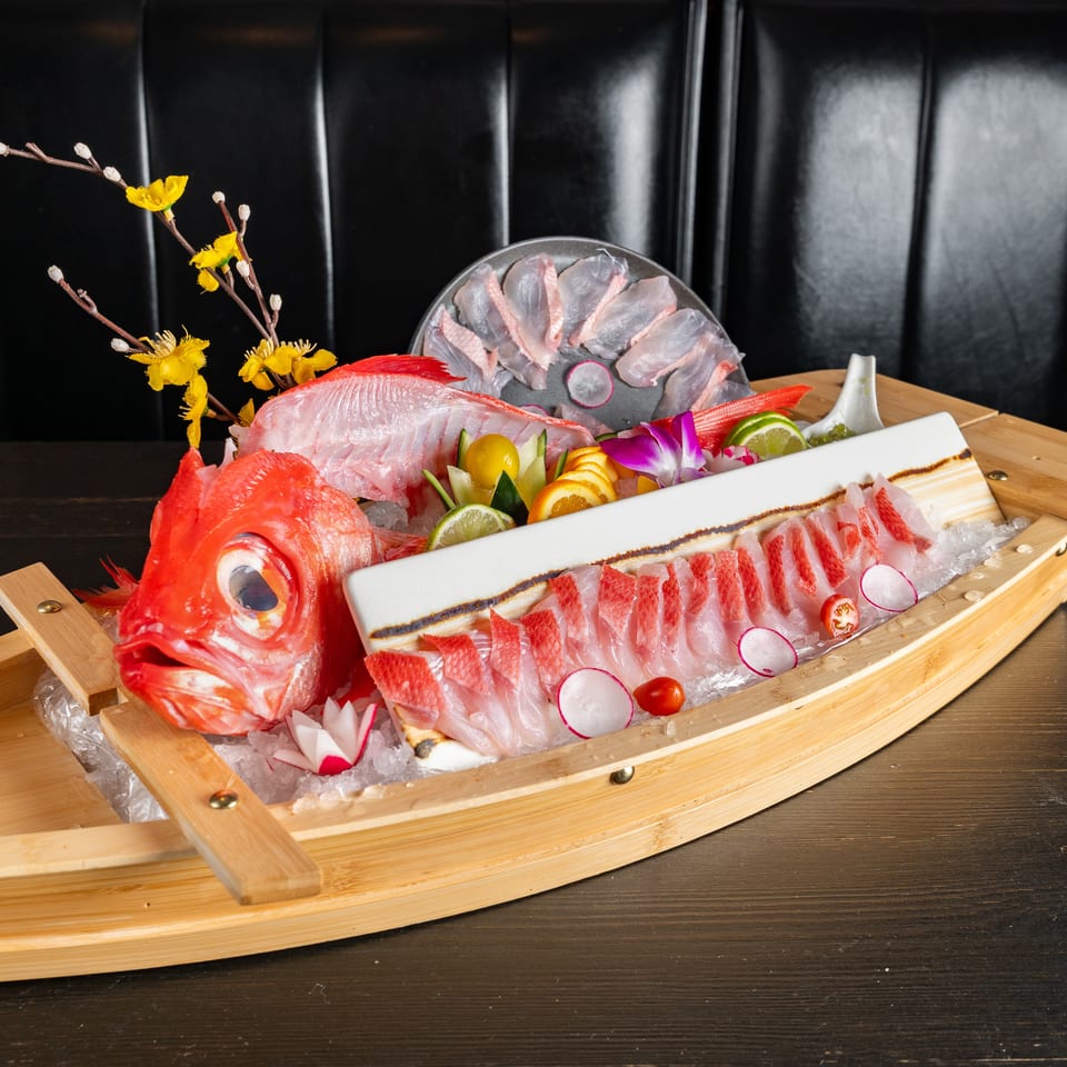 Large seafood sashimi platter arranged on ice in a wooden boat, garnished with citrus, vegetables, flowers, and a whole red fish.