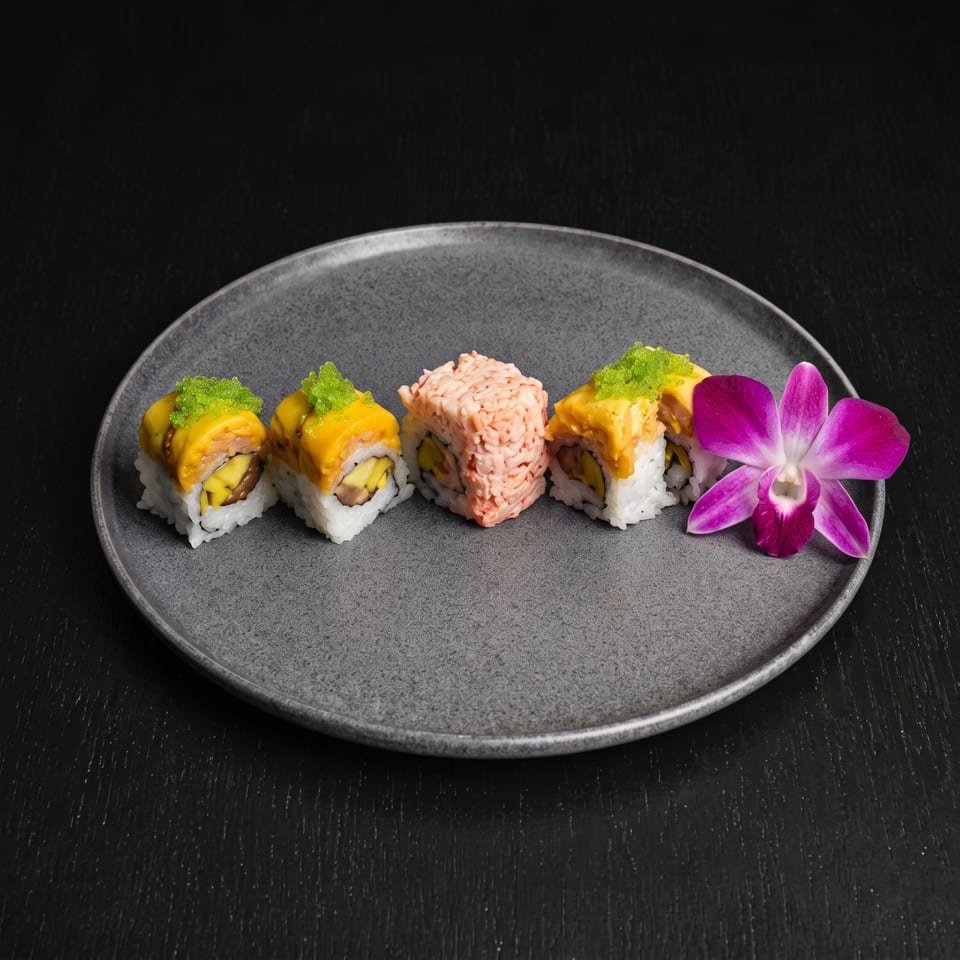 Assorted sushi rolls topped with yellow sauce and green garnish, plated with a pink flower on a dark ceramic plate.