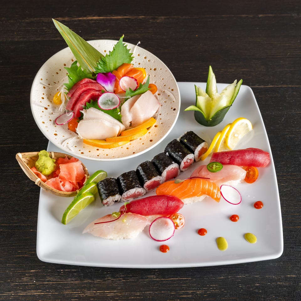 Assorted sushi and sashimi platter garnished with vegetables, lemon, lime, wasabi, and pickled ginger on a dark wooden table.
