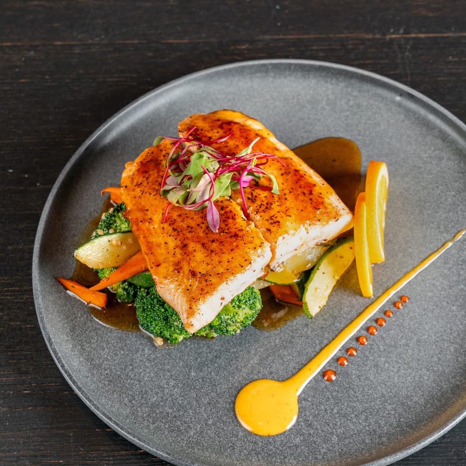 Seared white fish fillets with microgreens, roasted vegetables, orange slices, and sauce on a gray plate.