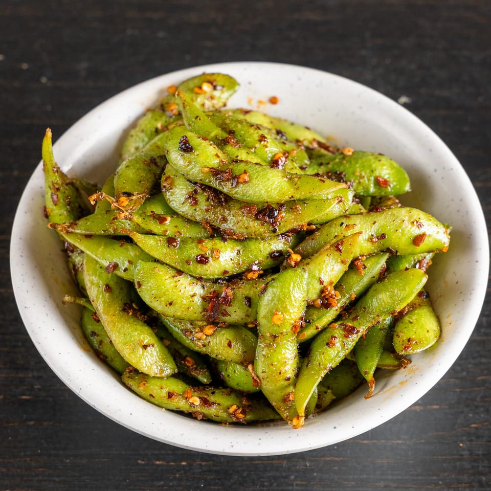 A bowl of green edamame pods seasoned with red pepper flakes and spices on a dark wooden table.
